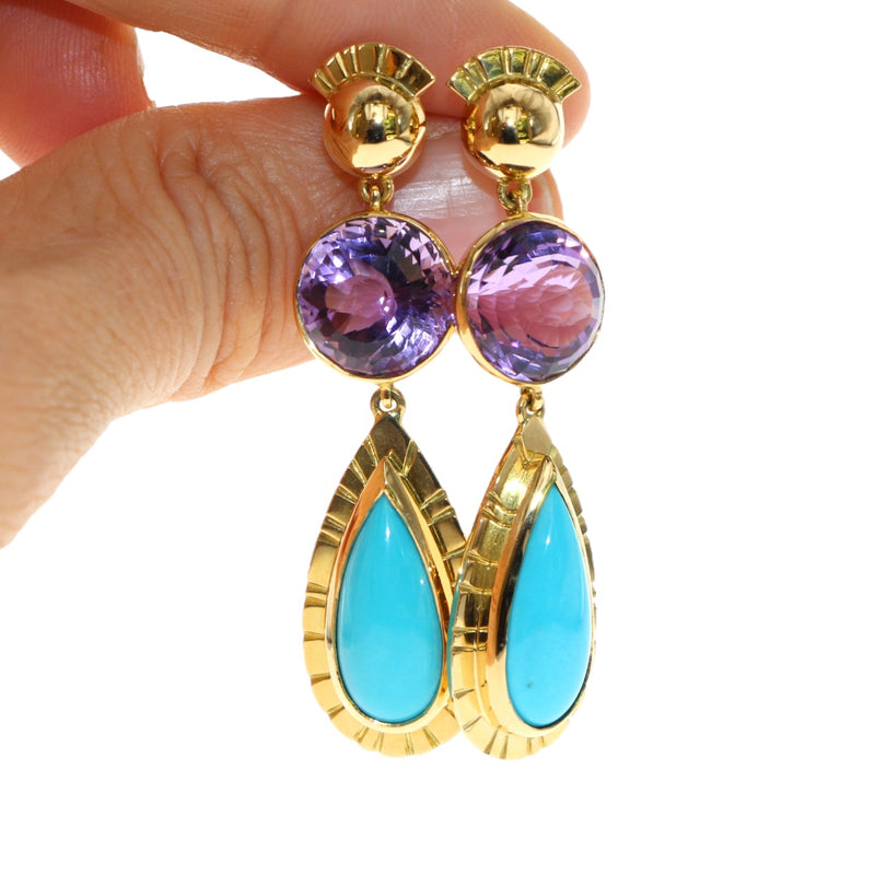 Gold earrings with purple and turquoise stones held by a hand against a white background